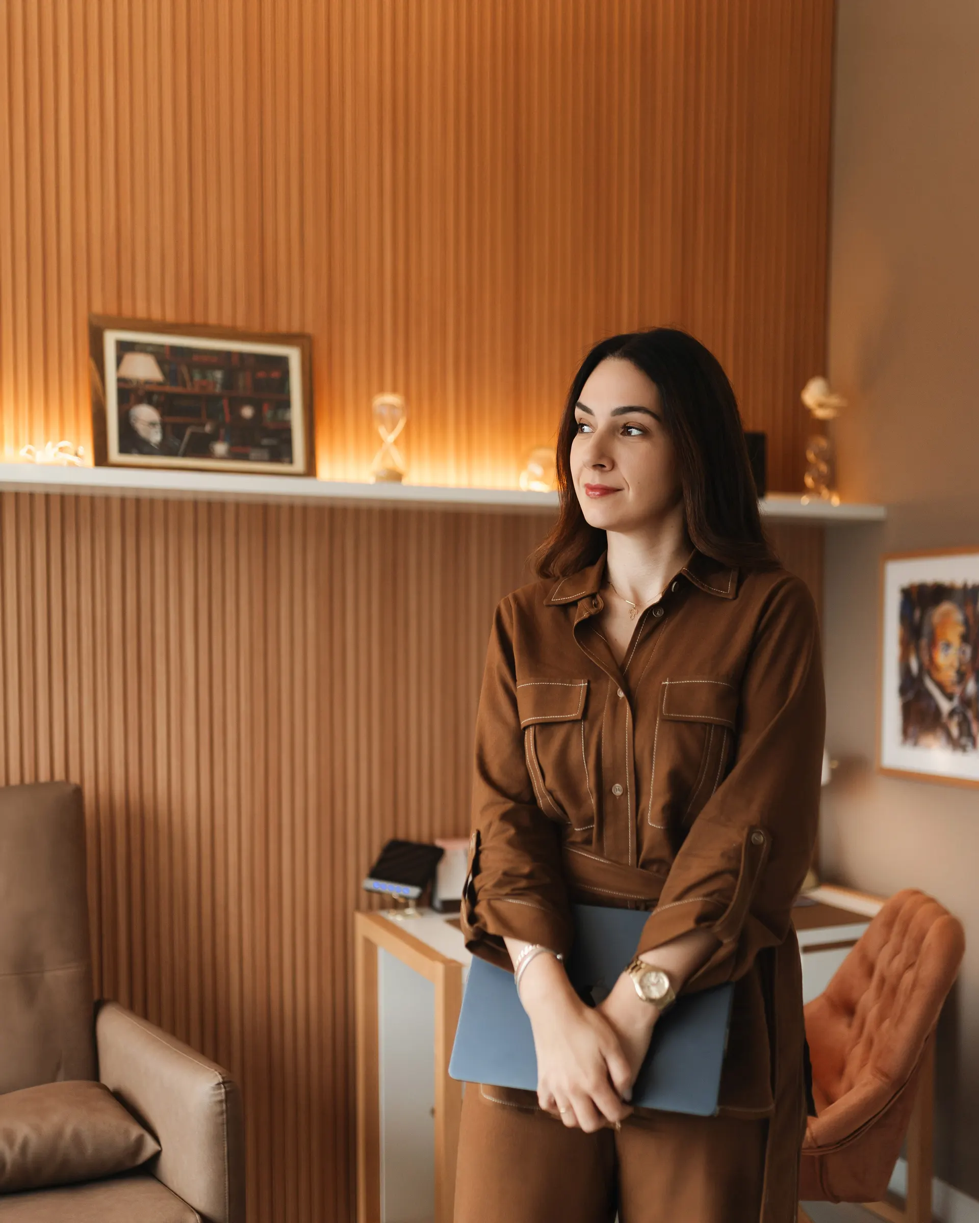 Gabriela Vargas em pé no consultório, segurando o notebook junto ao peito