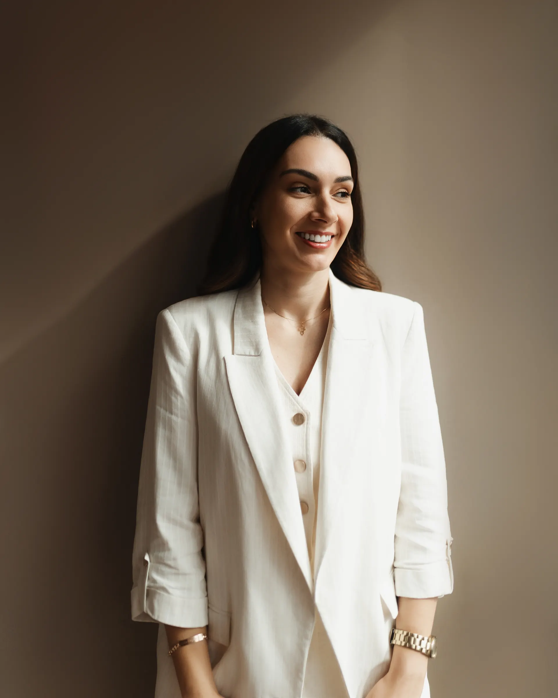Gabriela Vargas de blazer branco, apoiada na parede, sorrindo