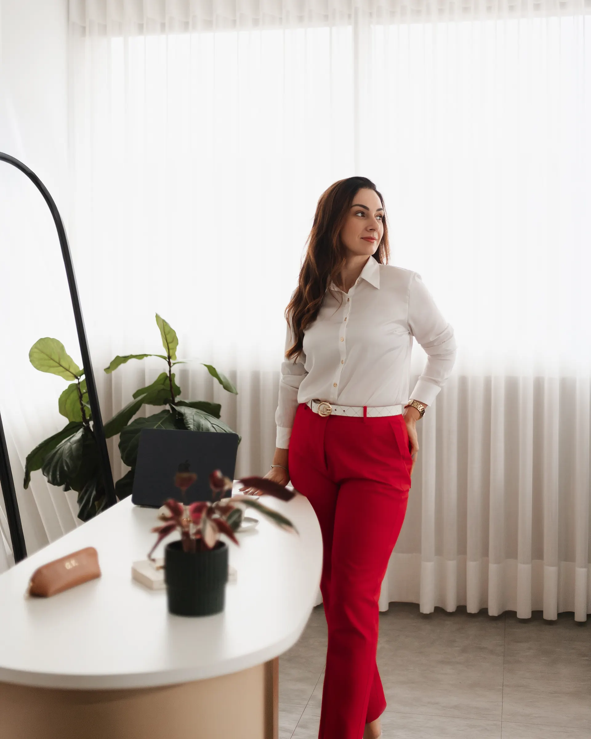 Gabriela Vargas de pé junto à janela do consultório, ao lado de uma planta