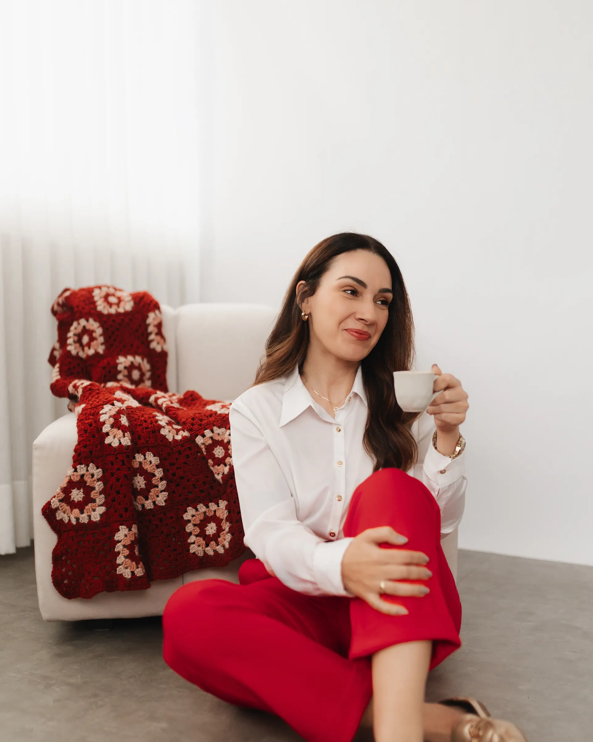 Gabriela Vargas sentada no chão do consultório, segurando uma xícara e sorrindo, com uma manta de crochê ao fundo
