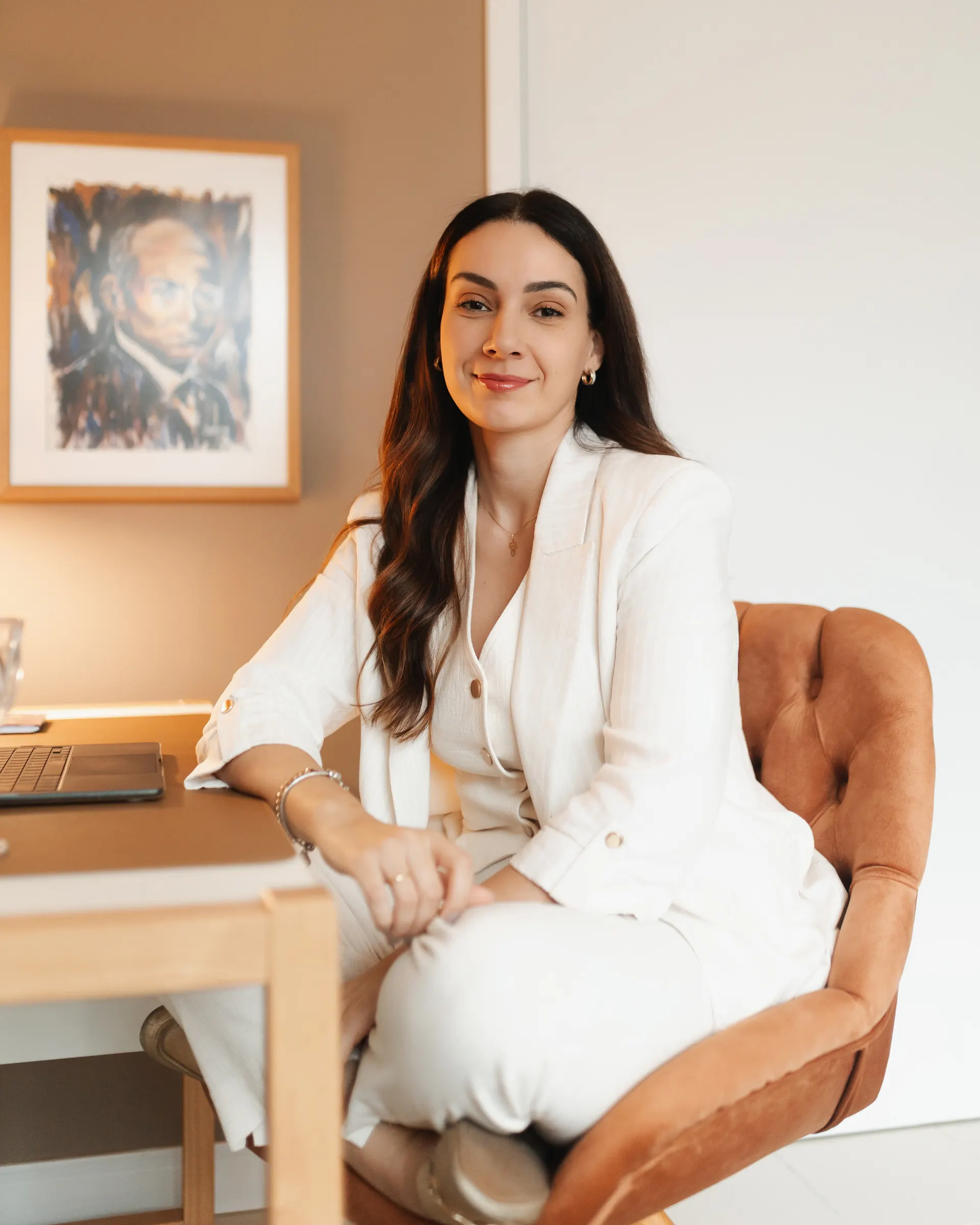 Gabriela Vargas de blazer branco, sentada à mesa do consultório, olhando para a câmera