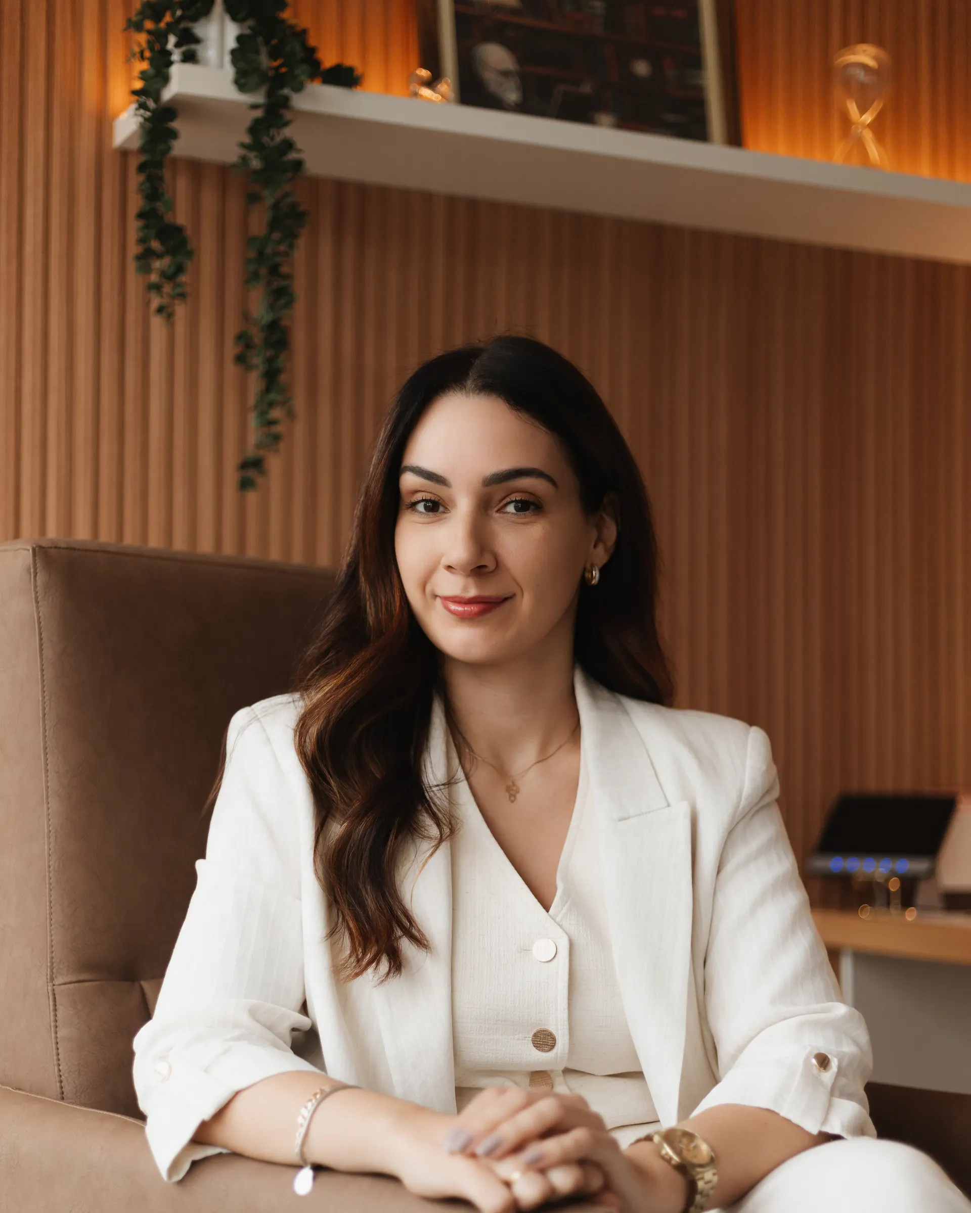Gabriela Vargas de blazer branco, sentada com as mãos cruzadas, olhando para a câmera